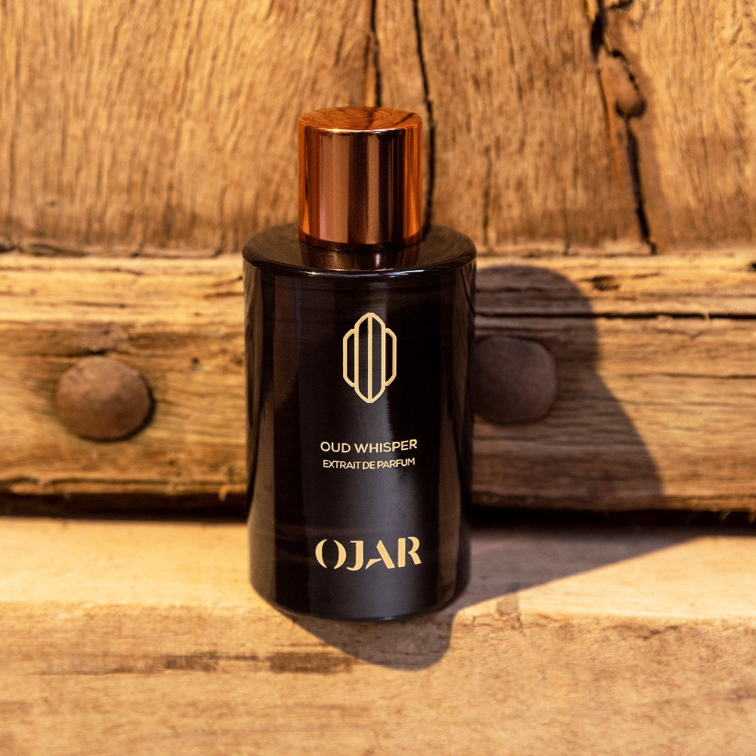 Black perfume bottle with gold cap labeled 'Oud Whisper' by 'Ojar' on a wooden surface.
